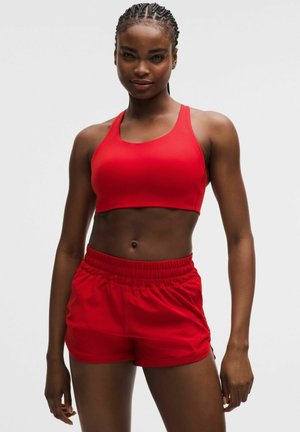 Atletische vrouw die een rode sportbeha en rode shorts met elastische tailleband draagt, staand met ontspannen armen tegen een effen achtergrond.
