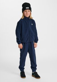 Mörkblå fleece-träningsdräkt med zip-up jacka och elastiska ärmmuddar, matchad med en svart mössa med en vit märkesetikett. Svarta sneakers med gröna detaljer.