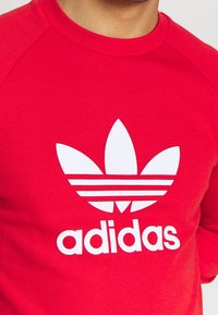 Červená mikina s bílým logem ve třech pruzích a textem "adidas". Má klasický kulatý výstřih a raglánové rukávy vyrobené z měkké látky.