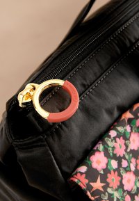 Černá taška s měkkou texturou, vybavená zlatým jezdcem zipu a červeným gumovým doplňkem. Na okraji je vidět látka s květinovým vzorem.
