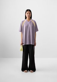Une jeune femme se tient contre un fond uni, portant une chemise oversized lavande, un pantalon ample noir, et tenant un petit sac jaune.