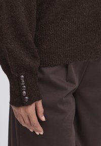 Maglione lavorato a maglia marrone con texture a coste e polsini con bottoni. Tessuto liscio con fibre visibili, abbinato a pantaloni scuri a vestibilità comoda.