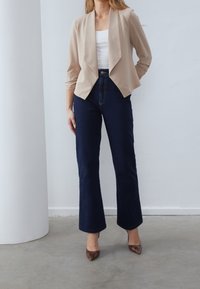 Beżowa krótka marynarka z drapowanym przodem, zestawiona z ciemnoniebieskimi szerokimi spodniami dżinsowymi. Brązowe buty na wysokim obcasie w wzór skóry węża dopełniają stylizację.