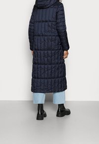 Abrigo acolchado largo en color azul marino con capucha, que cuenta con textura acolchada, bolsillos laterales y una longitud a medio muslo, usado sobre unos jeans cortos de color azul claro.