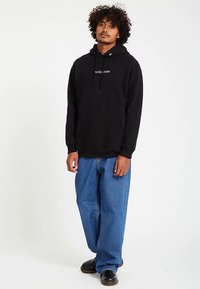 Sweatshirt preto com capuz, bolso frontal e cordões; apresenta o logótipo branco "VOLCOM". Combinado com jeans azuis largos e sapatos pretos.