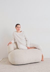 Frau in leichter gerippter Loungebekleidung, die barfuß auf einem großen, runden, cremefarbenen gepolsterten Sessel in einem minimalistischen Raum entspannt.