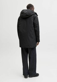 Črna vodoodporna parka s kapuco, značilna po dolgem, ravnem kroju in minimalističnem oblikovanju. V kombinaciji z temnimi širokimi hlačami in čevlji.