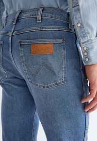 Közeli kép egy személyről, aki kék Wrangler farmert és hozzáillő farmer inget visel, amelyen márkás folt található a hátsó zsebén és ezüst kapcsos gombok az ujjon.