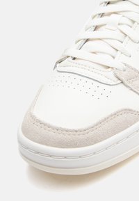 Sapatilha de couro branco com detalhes em camurça cinza, caixa do dedão perfurada, atacadores brancos planos e sola de borracha. Forma arredondada e detalhes costurados.