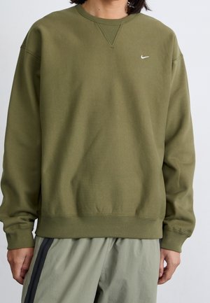Osoba ubrana w oliwkową bluzę Nike i jasne zielone spodnie z czarnymi paskami po bokach, stojąca na jednolitym tle.
