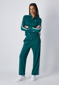Chándal en color teal con rayas laterales blancas, chaqueta con cremallera y pantalones con cintura elástica. Combinado con zapatillas blancas, tejido suave, diseño casual.
