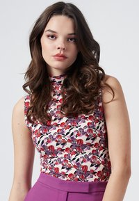 Ärmelloses Blumenshirt mit hoch geschlossenem Kragen in Creme, verziert mit roten und lila Blumen, kombiniert mit einfarbigen, lila, hochtaillierten Hosen.