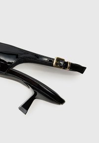 Črne lakirane slingback čevlje z visoko peto, z ozkim nosom in kvadratno peto. Ima zlat kovinski sponko na traku.