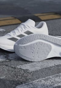 Chaussures de sport blanches avec des bandes grises, l'une montrant la semelle à motifs, placées sur un bitume fissuré près d'un trottoir jaune et noir.