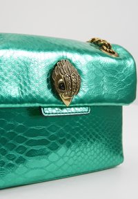 Borsa a mano in pelle di serpente verde con finitura metallica lucida, caratterizzata da una chiusura a forma di uccello dorato e una tracolla a catena. Superficie testurizzata con scaglie visibili.