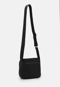 Černá crossbody taška s nastavitelným popruhem a klopovým uzávěrem, zobrazená zavěšená na hladkém bílém pozadí.