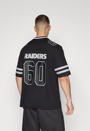 Man draagt een zwart Raiders-shirt met witte strepen en nummer 60 op de rug, staand tegen een effen lichte achtergrond.