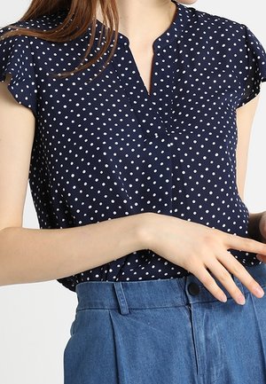 Femme portant un chemisier bleu marine à manches courtes avec des pois blancs et un pantalon bleu taille haute, une main reposant près de sa taille.