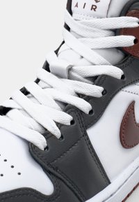 Närbild av en vit, grå och brun sneaker med texturerade vita snören som är trädde genom svarta öglor över en vadderad tunga.