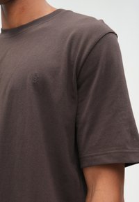 Brun bomullströja med en avslappnad passform, korta ärmar, rundad halsringning och en liten broderad logotyp på bröstet. Slät struktur.