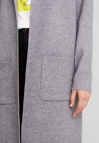 Šedý vlněný cardigan s dlouhými rukávy, otevřeným předním dílem a dvěma předními kapsami. Jemná textura a minimalistický design s útulným vzhledem.