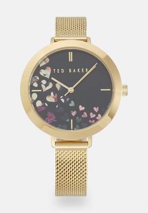 Gold mesh strap watch with black dial, gold hour markers, scattered colorful heart shapes, and "Ted Baker" logo at center.