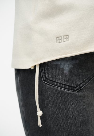 Μπεζ πουκάμισο με δύο μικρά λογότυπα σε σχήμα συν δίπλα στο τελείωμα και έναν κόμπο κορδονιού, φορεμένο πάνω από μαύρο τζιν με εμφανή πίσω τσέπη.