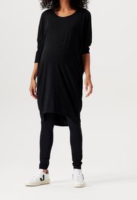 Vestido de maternidad negro, de corte suelto, con escote redondo y mangas tres cuartas, combinado con leggings negros y zapatillas blancas.