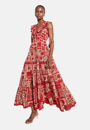 Kobieta w czerwonokremowej, bezrękawnej sukience maxi w stylu patchwork z falbanami, nosząca złote kolczyki koła i beżowe sandały na paskach.