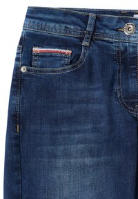 Gros plan sur la poche d'un jean en denim bleu foncé avec des coutures apparentes, des rivets, un bouton et une petite poche à monnaie avec un liseré rouge et blanc.