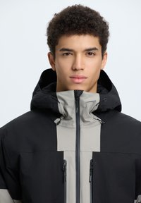 Giacca con cappuccio nera e grigia con cerniera frontale e due tasche con cerniera. Presenta un tessuto testurizzato e cordini regolabili sul cappuccio.