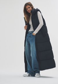 Hosszú fekete téli mellény kapucnival, steppelt mintával. Laza szabású kék farmerral és fehér sportcipővel párosítva.