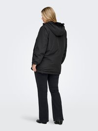 Woman with blonde hair wearing a black hooded puffer jacket, dark jeans, and black shoes, standing and facing away on a white background.