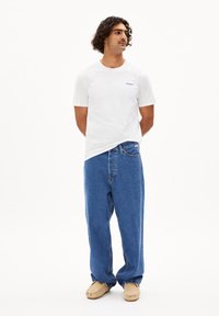 Witte t-shirt met korte mouwen en blauw logo, gecombineerd met losse blauwe jeans en beige instap schoenen. Eenvoudige, casual outfit.