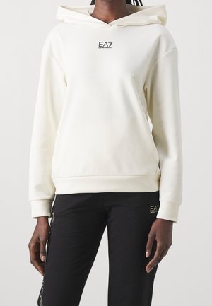 Personne portant un sweat à capuche EA7 Emporio Armani écru et un pantalon noir EA7 Emporio Armani sur un fond clair uni.
