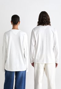 Witte longsleeveshirts met een V-hals aan de achterzijde, met een losse pasvorm. Het linker shirt is gecombineerd met blauwe jeans; het rechter shirt met witte broeken.