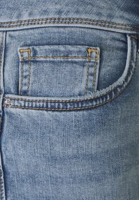 Hellblaue Jeans aus Denim mit einer Gesäßtasche, orangefarbenen Nähten und einem kleinen silbernen Nietendetail. Glatte Textur, ausgewaschener Look.