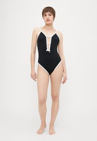 Maillot de bain une pièce noir à côtes avec des accents blancs, décolleté plongeant et détail de nœud à l'avant. Comprend des bretelles spaghetti et un design de jambe haute.