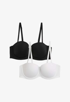 Kobieta w białym usztywnianym biustonoszu i pasujących majtkach z wysokim stanem, pozująca z uniesioną jedną ręką i spoglądająca w dół.