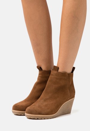 Bottines compensées en daim marron portées sur des jambes nues, avec languettes de tirage et semelles en caoutchouc texturé.