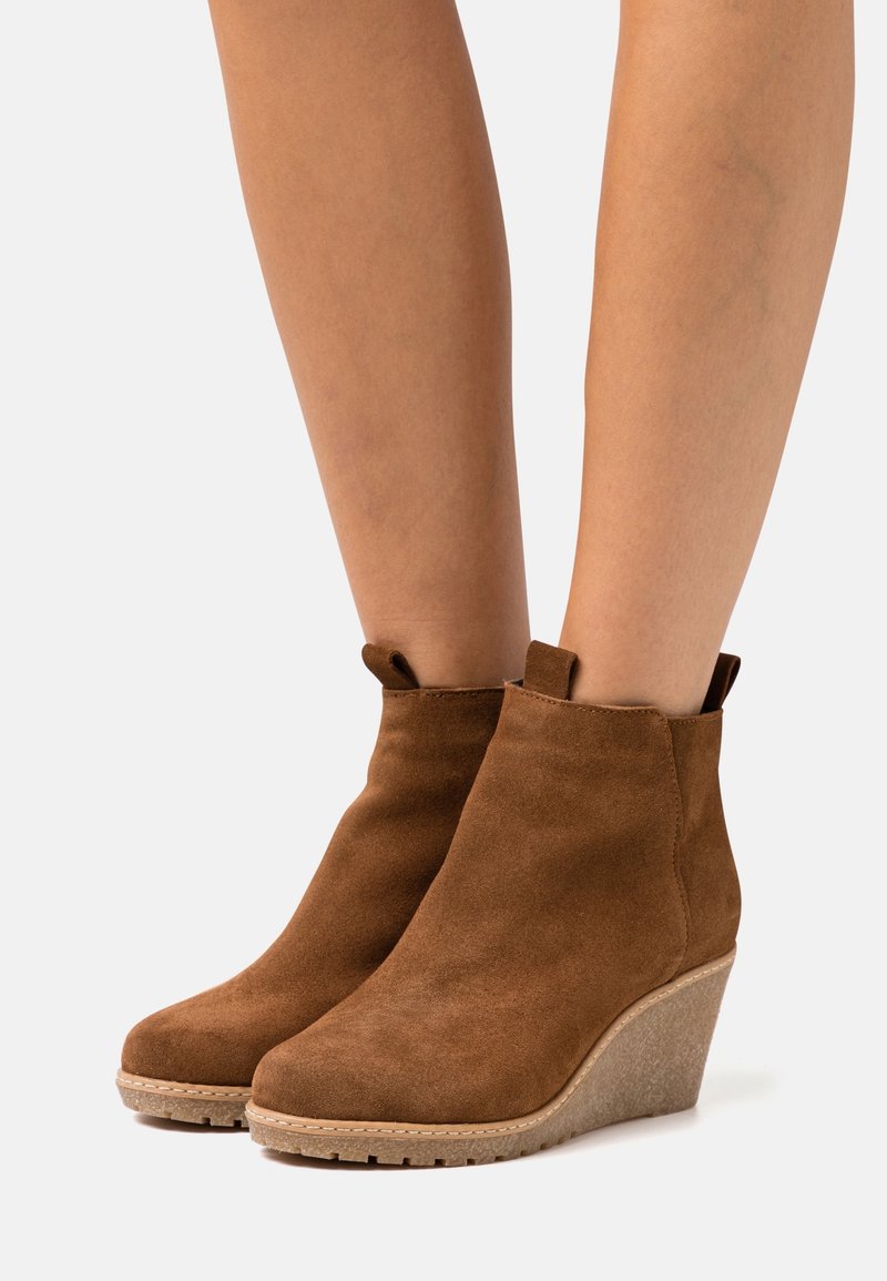 Bottines compensées en daim marron portées sur des jambes nues, avec languettes de tirage et semelles en caoutchouc texturé.