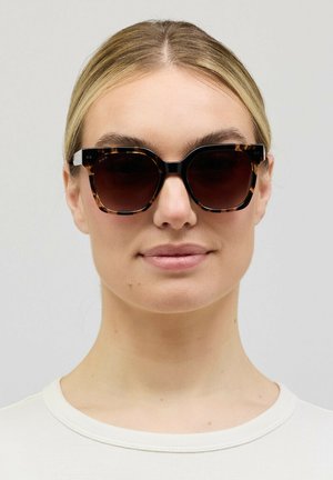 Vrouw met blond haar en grote zonnebril met schildpadmotief, gekleed in een wit shirt met ronde hals, die recht vooruit kijkt tegen een effen achtergrond.