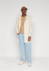 Beige vanntett jakke med hvit hette, beige T-skjorte, lyseblå jeans, kremfargede joggesko, og brun caps, stående foran en ensfarget bakgrunn.