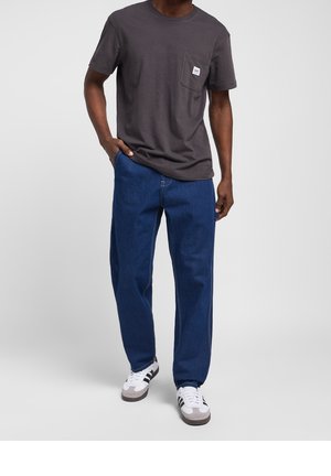 Homme portant un t-shirt gris foncé à poches, un jean bleu ample et des baskets blanches avec des bandes noires, debout devant un fond uni.