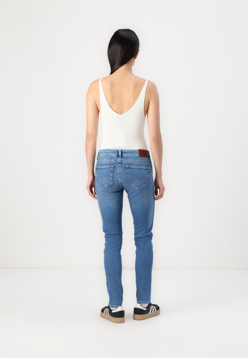 Weißes geripptes Tanktop, blaue Slim-Fit-Jeans mit einem braunen Lederpatch auf der Rückseite, schwarz-weiße Turnschuhe, von hinten betrachtet.