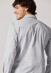 Bílé pánské košile s dlouhým rukávem a černým mřížkovým vzorem, s klasickým límcem a uvolněným střihem, se zřasením na zadní straně.