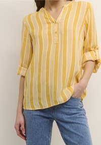 Gulrandig skjorta med notchkrage, knäppning framtill och uppvikta ärmar, tillverkad av lätt tyg, ihop med blå jeans.