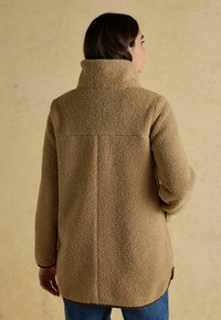 Kreemjas fliisjakke kõrge kraega, tekstuurse pinna ja sirge lõikega. Omab nähtavat õmblust seljaosas ja kõverat serva.