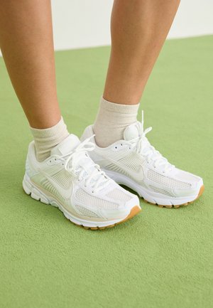 Legs wearing cream-colored ankle socks and white Nike running shoes standing on light green textured floor.