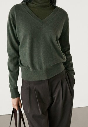 Person wearing a dark green V-neck sweater over a matching turtleneck, dark pleated pants, holding brown bag handles in left hand.